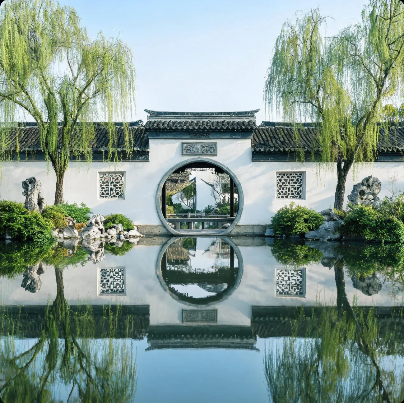 白墙圆门与两侧垂柳临水而立，墙面、树木和门洞的倒影映在池中。